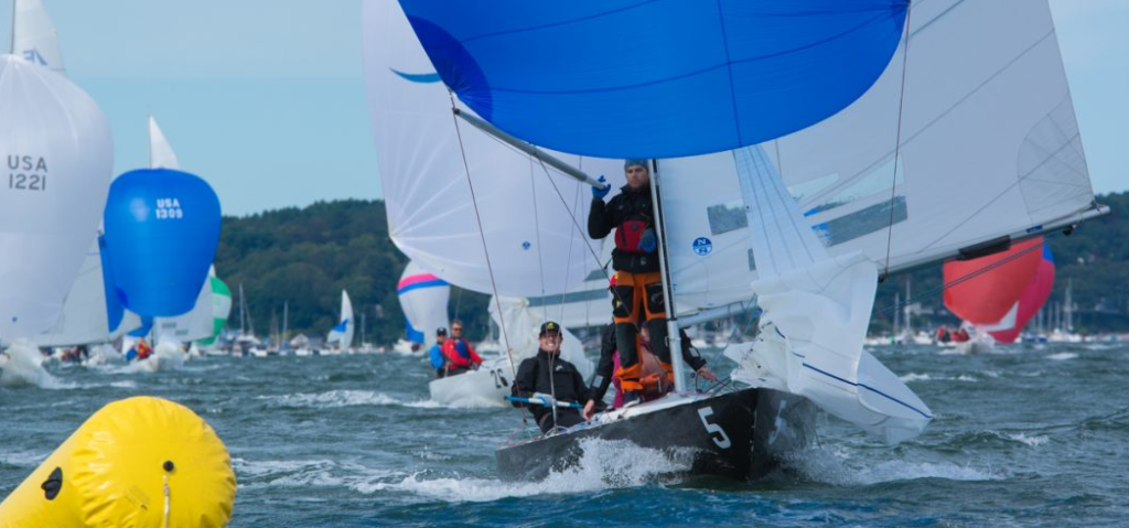 Sailboats with colorful spinnakers race on choppy water. A yellow buoy floats in the foreground, and two sailors in life jackets steer boat number 5. Trees and more sailboats are visible in the background.