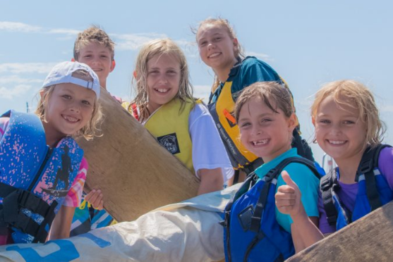 Six smiling children wearing colorful life jackets pose together outdoors on a sunny day, holding onto wooden boards and sails, suggesting they're participating in a water or boating activity. One child gives a thumbs up.