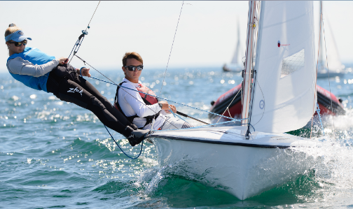 Two people sailing a small white sailboat on open water. One person leans back over the edge, supported by harness lines, while the other steers. They both wear sunglasses and athletic gear. Waves splash around the boat.
