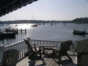 A sunlit deck overlooking a calm harbor with several boats anchored in the water; wooden chairs and a table sit on the deck, and sunlight reflects off the water's surface.