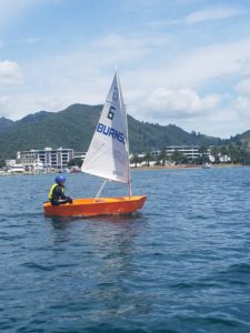 A person wearing a helmet and life jacket sails a small orange dinghy with a white sail on calm blue water, with buildings, palm trees, and green hills in the background under a partly cloudy sky.