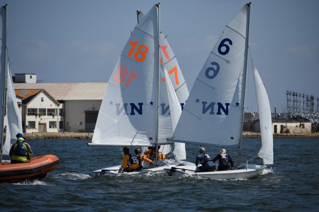 Three small sailboats with numbers 18, 17, and 6 on their sails are racing closely on the water near an industrial shoreline, with people in life jackets and a safety boat visible on the left.