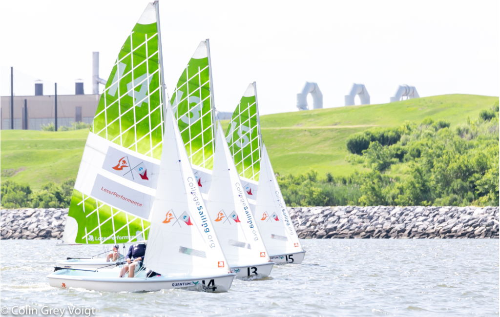 Three small sailboats with green and white sails race on the water near a rocky shoreline, with green hills and industrial buildings visible in the background.