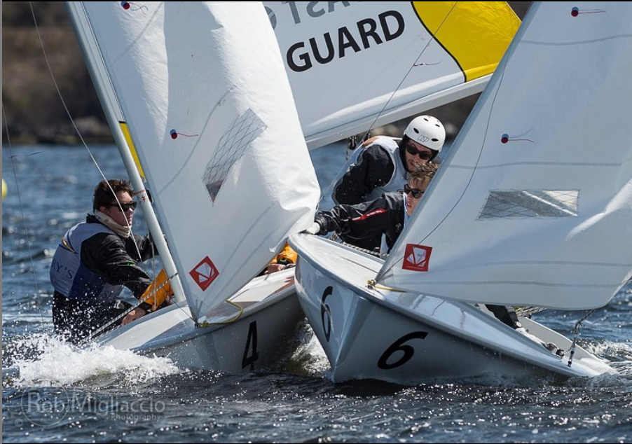 Two small sailboats with crews race closely in the water, their sails nearly touching. Four people are visible, wearing life vests and helmets, intensely focused as the boats compete.