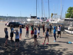 A group of teenagers stands in a circle on a sunny marina lot, stretching with hands behind their heads. Sailboats and cars are parked in the background near a chain-link fence.