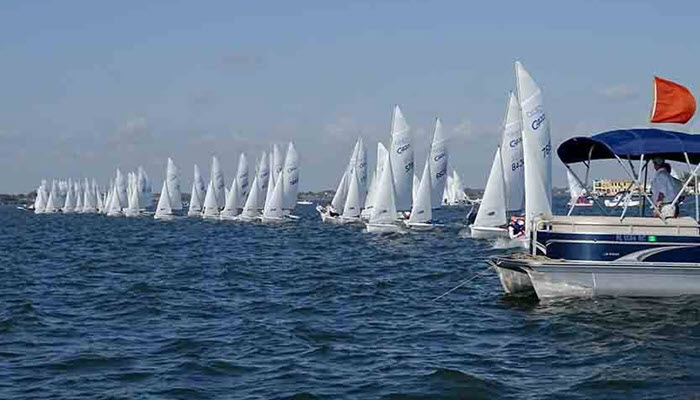 A fleet of white sailboats lined up on the water, ready to race under a clear blue sky. A motorboat with a red flag is in the foreground on the right side of the image.