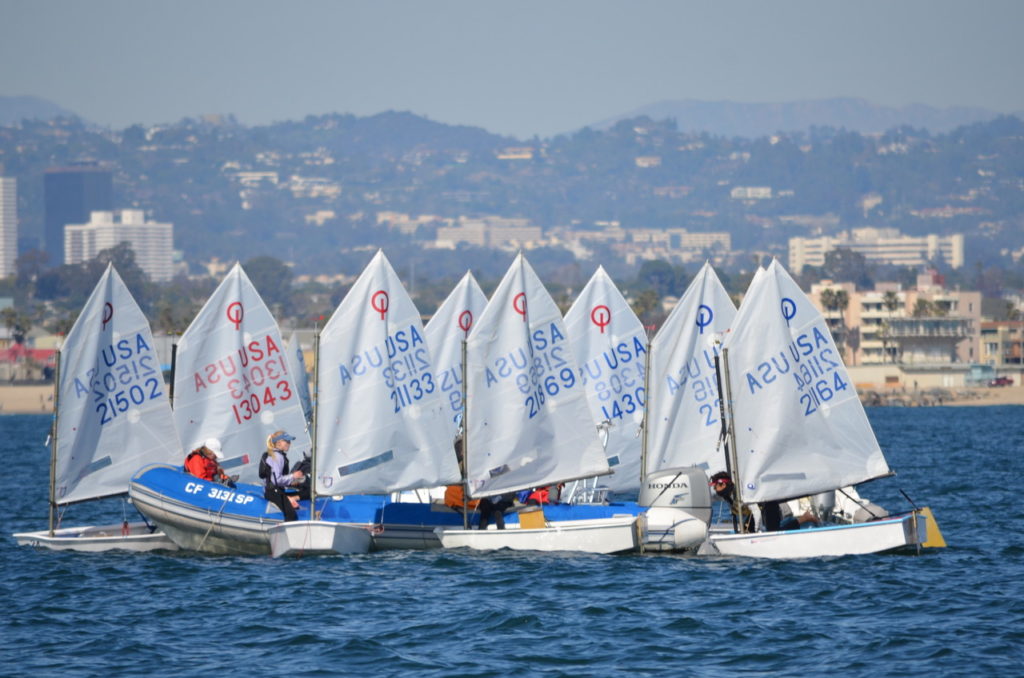A group of young sailors in small sailboats with white sails compete close together on the water near a city shoreline, with buildings and hills visible in the background.