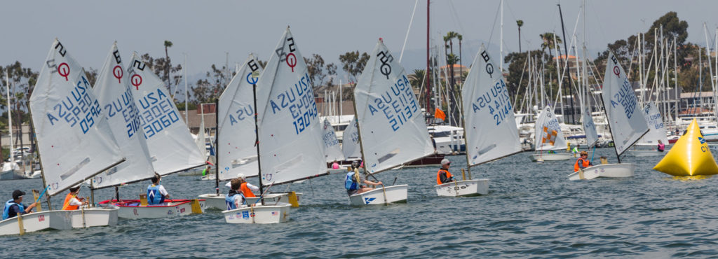 A group of young sailors racing small sailboats on a marina, passing a yellow buoy with docks, boats, and palm trees in the background under a clear sky.