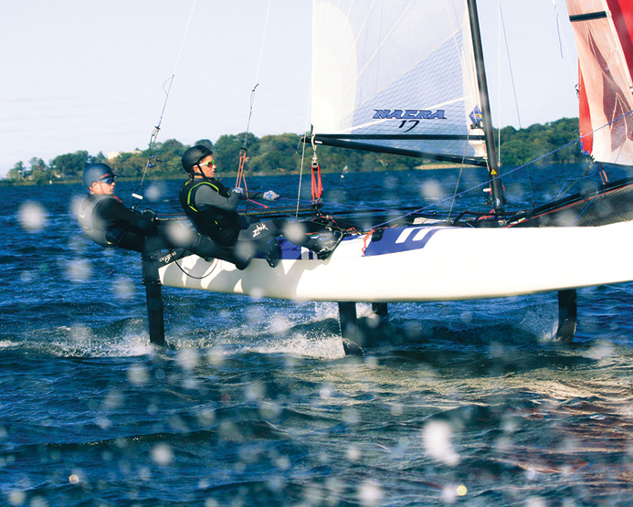 Two people in wetsuits and helmets sail a hydrofoil catamaran across a lake, leaning back to balance the boat. Water splashes in the foreground and trees line the distant shore under a clear sky.