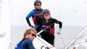 Two sailors wearing helmets and MIT sailing gear stand on a small sailboat, focused on maneuvering. Another person in the foreground is slightly out of focus. The background shows open water under a cloudy sky.