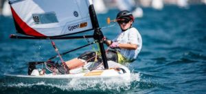 A young sailor wearing a helmet and sunglasses steers a small sailboat on a sunny, choppy lake, leaning back while gripping the tiller and mainsheet. Other sailboats are visible in the blurry background.