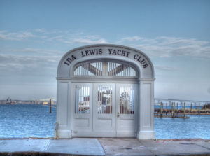 A white, arched entrance gate to the Ida Lewis Yacht Club stands by the waterfront, with a marina, docks, and a bridge visible in the background under a clear blue sky.