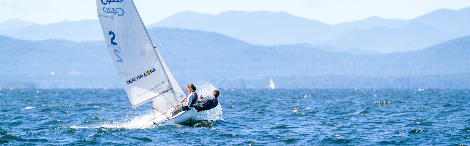 Two people sailing a small sailboat on a choppy lake, leaning to balance the boat, with distant mountains and a clear sky in the background.