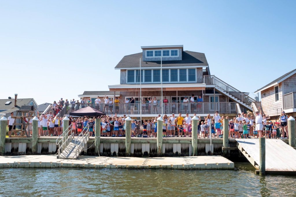 A large group of people gather on a dock in front of a multi-story beach house, waving and smiling toward the camera under a clear blue sky.