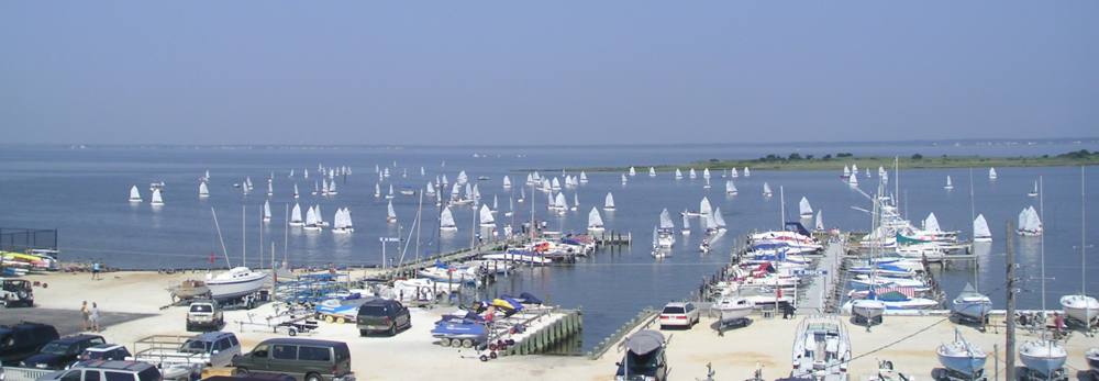 A marina filled with parked cars, boats, and sailboats, with numerous small sailboats scattered across a calm blue bay under a clear sky.