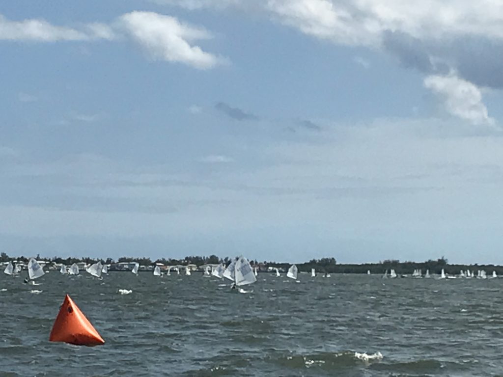 Many small sailboats with white sails are sailing on choppy water under a partly cloudy sky. An orange triangular buoy floats in the foreground, and a distant shoreline is visible in the background.
