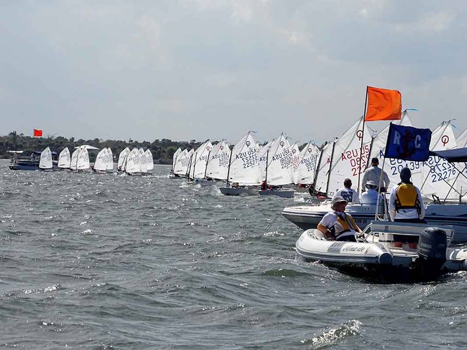 A fleet of small sailboats with white sails races on a choppy body of water, while people in a motorboat with flags observe and coordinate the event. Trees and a distant shoreline are visible in the background.
