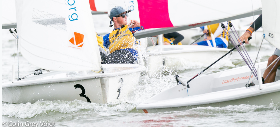 Several sailors compete in small sailboats on choppy water, wearing life vests and caps. Water splashes around the boats, with the number 3 visible on one hull. Bright sails and intense action are prominent.