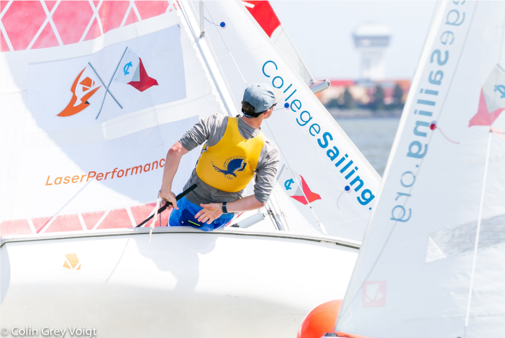 A sailor wearing a yellow life jacket and cap steers a sailboat labeled "College Sailing" and "LaserPerformance" during a bright, competitive sailing event.