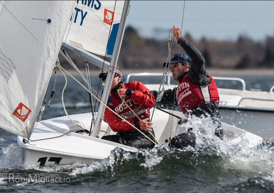 Two Boston University sailors wearing red life vests compete in a sailing race, maneuvering their boat through choppy water with visible splashes and focus. A safety boat is seen in the background.