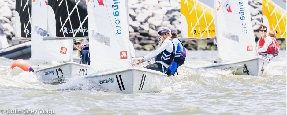 A group of young sailors competes in small sailboats on the water, wearing life jackets and sunglasses. Yellow and white sails with “Sailing.org” branding are visible, with a rocky shoreline in the background.