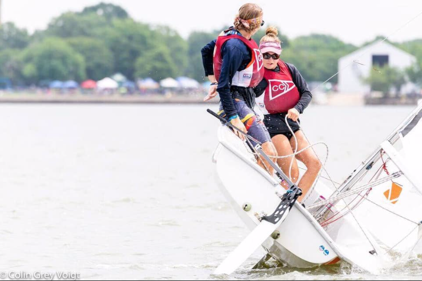 Two sailors in life vests and team uniforms balance on the edge of a small sailboat, leaning out as the boat tilts in the water during a regatta, with trees and tents visible in the background.