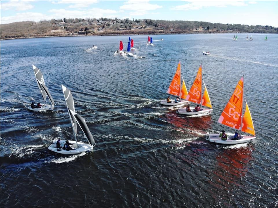 Several small sailboats with orange and black sails race on a wide river, leaving trails in the water. More boats are visible in the distance, with an overcast sky and tree-lined shore in the background.