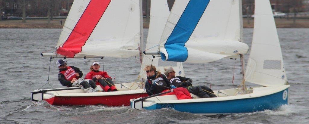 Two small sailboats race on choppy water, each with two people onboard. The left boat is red with a white and red sail, while the right boat is blue with a white and blue sail. Trees and buildings are visible in the background.