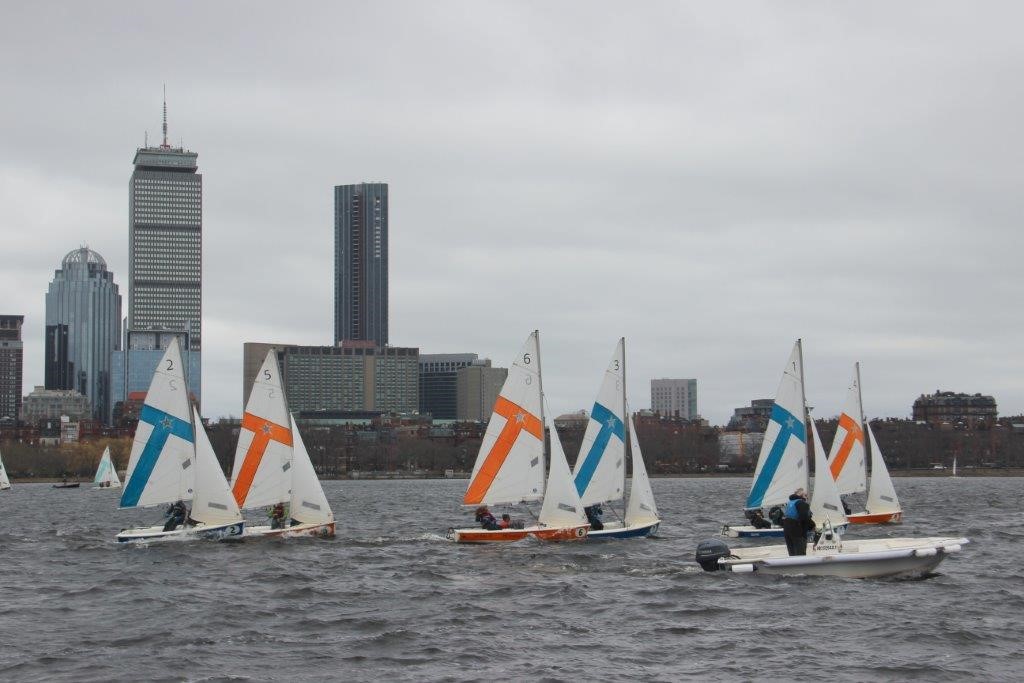 Small sailboats with orange and blue sails race on a river with a city skyline and tall buildings in the background under a cloudy sky. A motorboat is nearby on the water.
