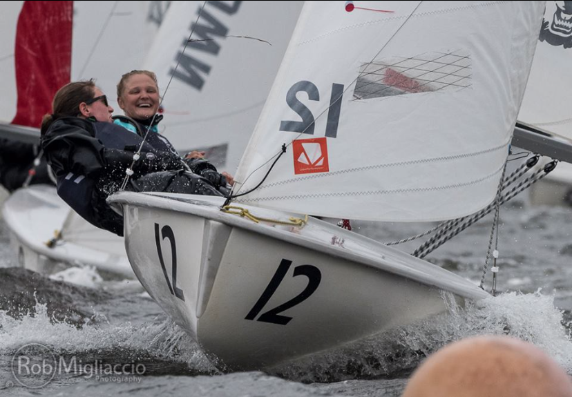 Two sailors smile and lean back in a small sailboat marked "12," racing in choppy water with other sailboats visible in the background. The image captures motion and excitement during the sailing competition.