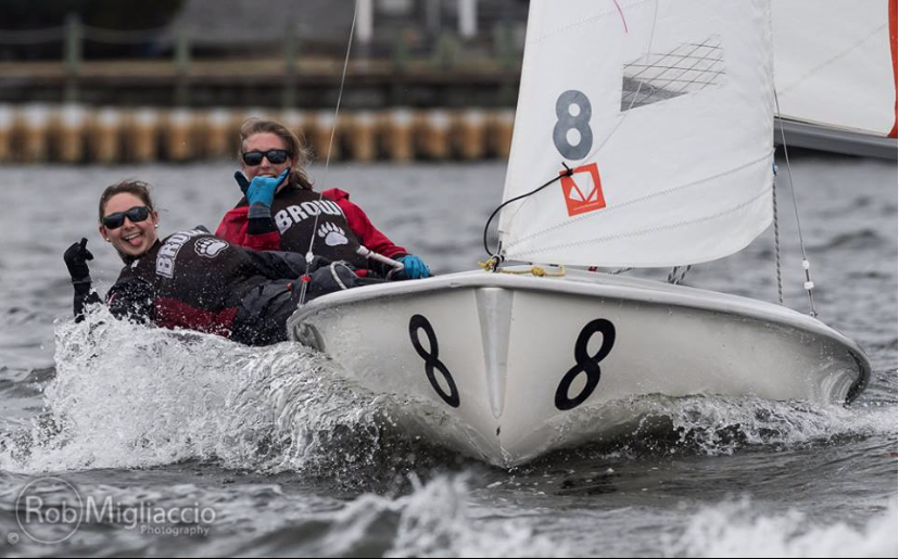 Two smiling sailors in life jackets ride a small sailboat numbered '8' across choppy water, creating a splash. Both gesture happily towards the camera. The background shows a dock and blurred shoreline.