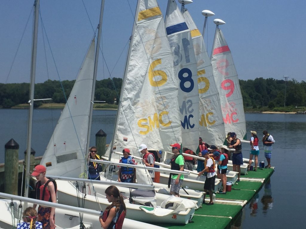 A group of people, many wearing life jackets, stand on a dock next to several small sailboats with numbered sails on a calm lake, preparing for sailing on a sunny day. Trees and grass are visible in the background.