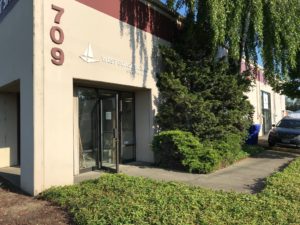 A beige building with the address number 709, glass entrance doors, and a West Coast Sailing sign. A tree and bushes are near the entrance, and a parked car is visible on the right.