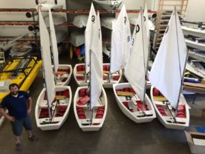 A man stands smiling in a warehouse next to six small sailboats with white sails and red seats, arranged in two rows. Various other boats and equipment are stored on racks in the background.
