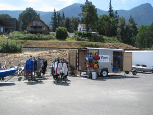 A mobile shop trailer with racks of jackets and clothing set up outside in a parking lot, with houses on a hill and forested mountains in the background under a clear sky.