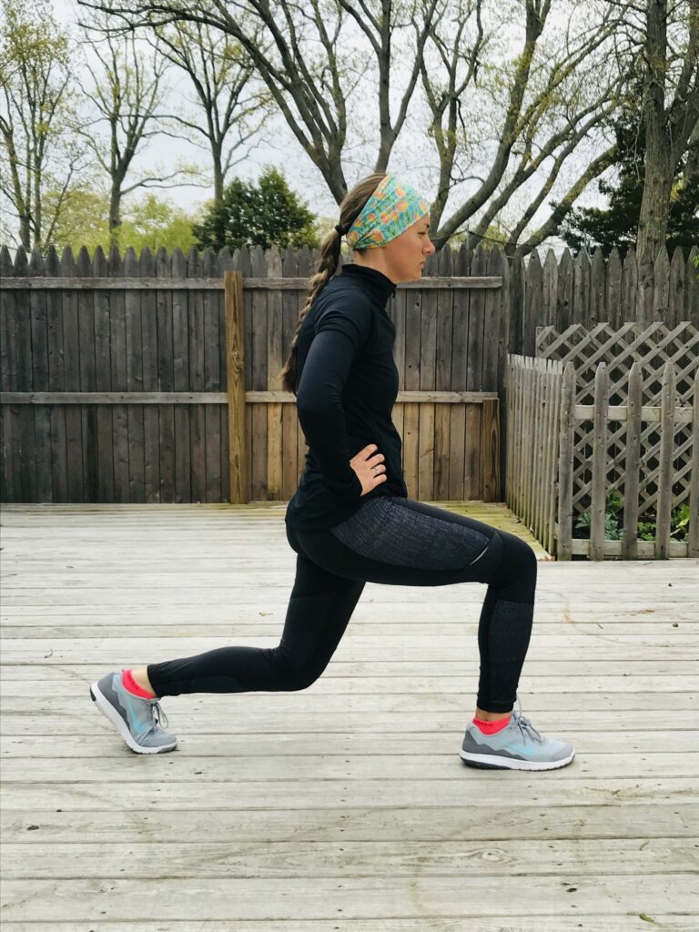 A woman wearing athletic clothes and a headband performs a forward lunge on a wooden deck outdoors, with hands on hips and trees visible in the background.