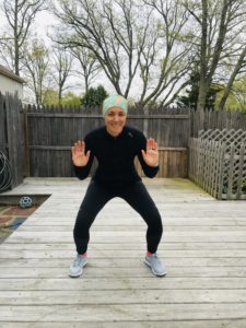 A person in athletic wear and a colorful headband is smiling and holding their hands up while squatting on a wooden deck outdoors, with leafless trees and a wooden fence in the background.