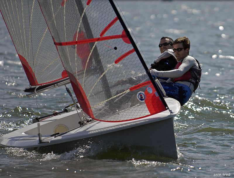 Two people in life jackets sail a small racing dinghy with red and white sails on a sunny day, leaning out over the side as the boat tilts on the water.