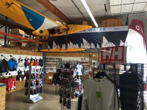 Interior of a water sports store with kayaks hanging from the ceiling, sailboats pictured on a banner, clothing racks with wetsuits and shirts, and shelves displaying various accessories and equipment.