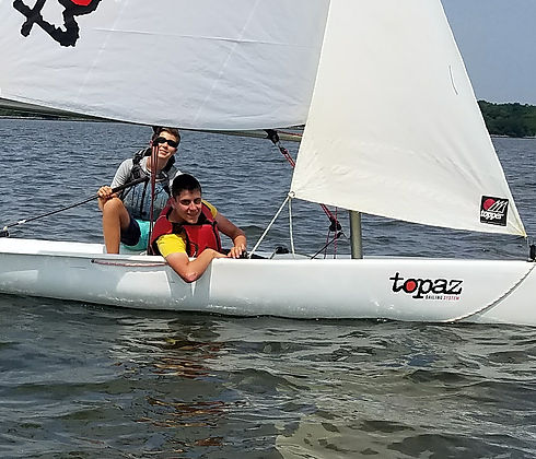 Two young people are sailing a small white sailboat labeled "topaz" on a calm body of water, both wearing life jackets and smiling. Trees and shoreline are visible in the background.