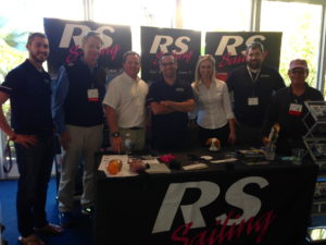 Seven people stand and smile behind an RS Sailing promotional table at an indoor event, with RS Sailing banners in the background and various items on the table, including brochures and small display items.