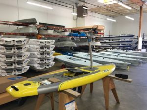 A yellow paddleboard with a handle is displayed on sawhorses in a warehouse, surrounded by stacked white and blue boats, boat parts, and shelves under bright overhead lighting.