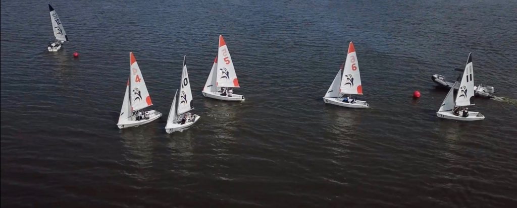 Five small sailboats with red and white sails race closely together on dark blue water, accompanied by a motorboat on the right side of the image. A red buoy floats in the distance.
