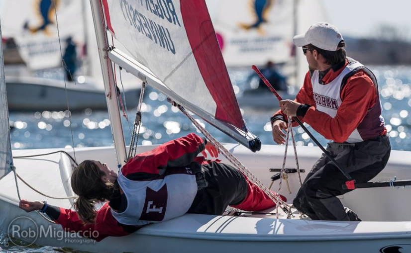 Two sailors in red gear are sailing a small boat. One sailor leans back over the edge, while the other sits upright, holding the tiller. Both wear life jackets and the water is visible in the background.