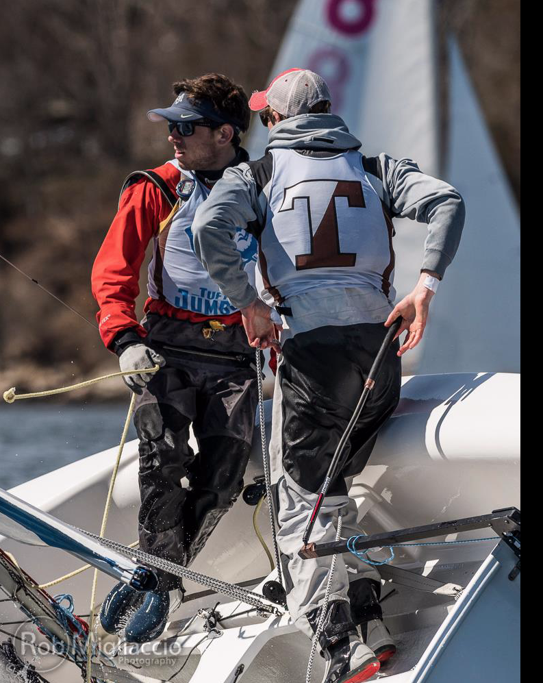 Two sailors in gear stand on a sailboat during a race, looking in different directions. One wears a red jacket and sunglasses, the other a gray hoodie with a large “T” on the back. Water and blurred trees are visible in the background.