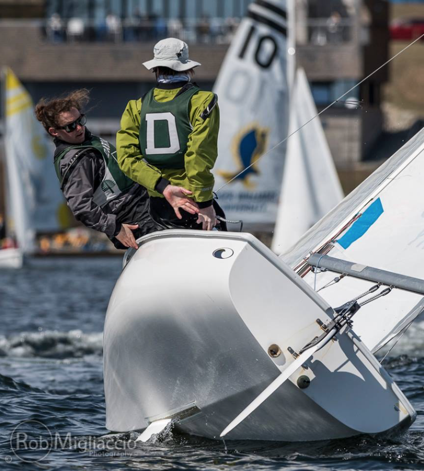 Two sailors lean out of a small white sailboat, navigating a turn in a race. Other sailboats and a building are visible in the background on the water. Both sailors wear life jackets and hats for sun protection.