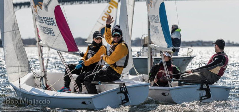 Two small sailboats with teams of young people on the water. One person in the front boat waves and smiles at the camera while others steer and adjust sails. The sun shines on the water, and a bridge is visible in the background.