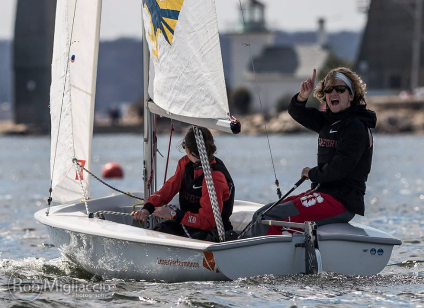 Two people sail a small white boat on open water. One person, wearing sunglasses, points upward and smiles, while the other steers. Lighthouse structures and a buoy are visible in the background.