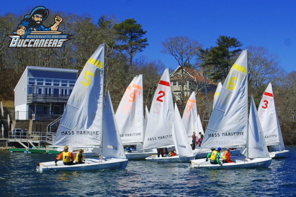 Several sailboats with “Mass Maritime” on the sails compete near a shoreline with buildings and trees. The Massachusetts Maritime Buccaneers logo is in the top left corner. Sailors wear life jackets under a clear blue sky.