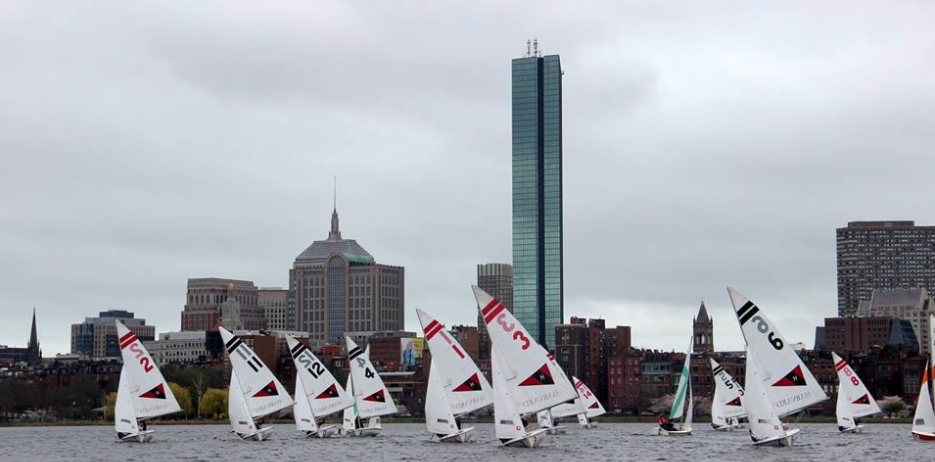 Several small sailboats with numbered sails race on a river, with a city skyline and tall glass skyscraper in the background under a cloudy sky.